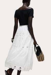 Svart off-shoulder topp tillsammans med en vit, flerskikts maxi kjol med spetsdetaljer, hålls med ena handen; accessoriserad med en vävd väska.