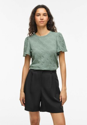 Femme portant un haut vert texturé à manches courtes associé à un short noir taille haute sur mesure, debout devant un fond blanc uni.