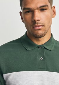 Zöld és szürke csíkos galléros póló texturált anyagból, gallérral, két gombbal és egy vízszintes szürke csíkkal a mellrészen.