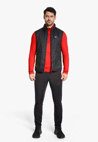Schwarze gepolsterte Weste über einem roten Langarmshirt, kombiniert mit schwarzen Hosen und Schuhen. Mit einem Reißverschluss vorne und einem Logo auf der Weste.