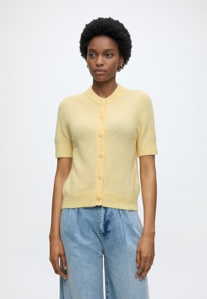 Femme portant un cardigan court boutonné jaune clair et un jean bleu clair ample, debout devant un fond blanc uni.