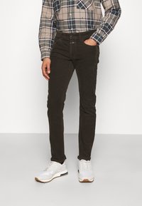 Pantalon en velours côtelé marron à coupe slim, avec une fermeture à boutons à l'avant et des poches, assorti à des baskets blanches et une chemise à carreaux.
