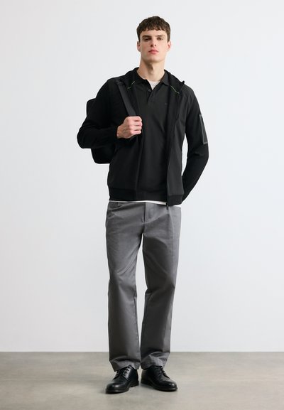 Jeune homme portant une veste noire zippée, un polo noir, un pantalon gris, des chaussures noires, portant un sac à dos noir sur une épaule, debout contre un mur blanc.