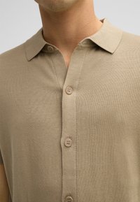 Beige polo shirt met korte mouwen van zacht gebreid materiaal, voorzien van een kraag en drie knopen aan de voorkant voor het sluiten.