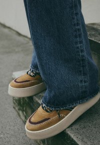 Braune Wildlederschuhe mit kastanienbraunen Akzenten, gesteppten Details und schwarzen Schnürsenkeln; getragen mit blauen Jeans im weit geschnittenen Stil.