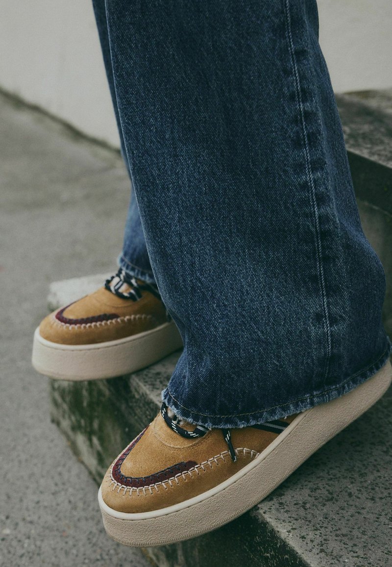 Braune Wildlederschuhe mit kastanienbraunen Akzenten, gesteppten Details und schwarzen Schnürsenkeln; getragen mit blauen Jeans im weit geschnittenen Stil.