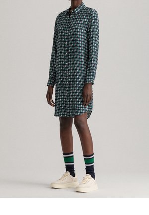 Osoba ubrana w granatową sukienkę koszulową z długim rękawem w wzory, zielone i białe skarpetki oraz białe trampki, stojąca na jednolitym tle.