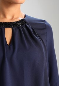 Blouse bleu marine avec un décolleté en trou de serrure, des détails froncés et des accents perlés autour du col. Confectionnée dans un tissu lisse et fluide.
