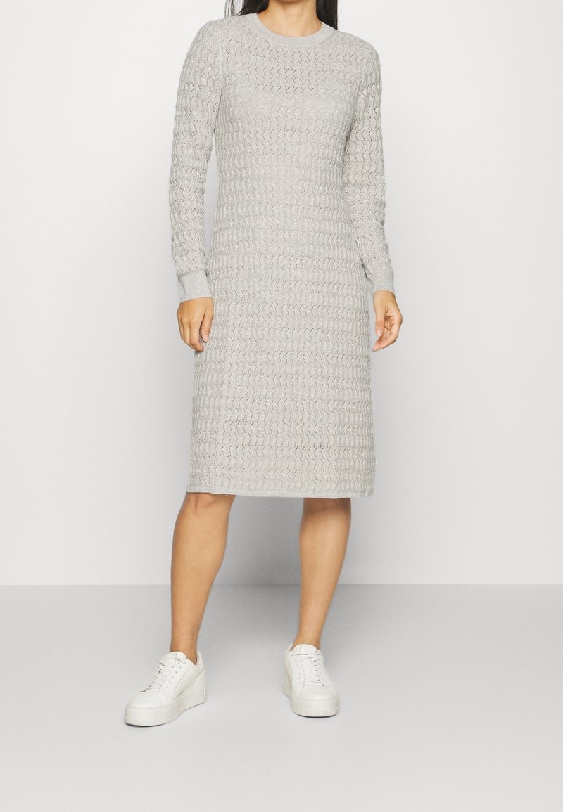 Femme portant une robe en maille texturée gris clair, longueur genou et manches longues, associée à des baskets blanches à plateforme, debout devant un fond uni.