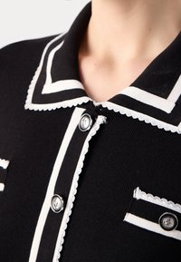 Vêtement en maille noire avec bordure festonnée blanche, rayures blanches sur le col et les poches, et boutons argentés près de l'encolure et de la poche poitrine.