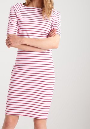 Kvinde iført en hvid knælængde kjole med røde vandrette striber, stående med armene krydset mod en ensfarvet baggrund.