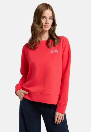 Rotes Sweatshirt mit überschnittenen Schultern, rundem Halsausschnitt und weißem Logo auf der Brust. Weiche Textur, kombiniert mit marineblauen Hosen.