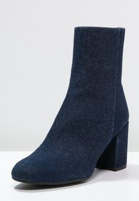 Bottine en denim avec un haut talon bloc, un bout pointu et une texture lisse. Présente une couleur bleu marine profond et une silhouette élégante.