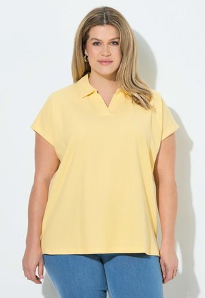 Kobieta o jasnobrązowych włosach, ubrana w żółtą koszulkę polo z krótkim rękawem i niebieskie dżinsy, stojąca na białym tle.