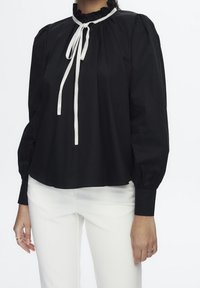 Zwarte blouse van soepele stof, met een gerimpelde halslijn, lange mouwen en witte strikjes aan de voorkant. Draag met witte broek.