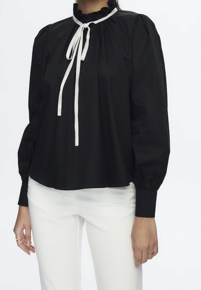 Zwarte blouse van soepele stof, met een gerimpelde halslijn, lange mouwen en witte strikjes aan de voorkant. Draag met witte broek.