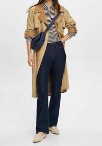 Cappotto trench beige, maglietta a righe bianche e nere, jeans svasati blu scuro, mocassini beige e borsa a tracolla blu navy. Texture morbide.