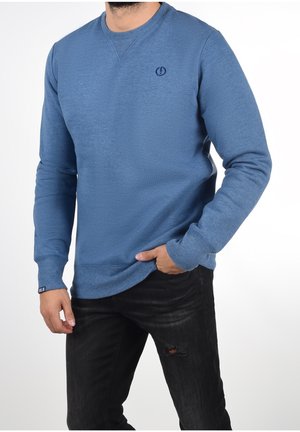 Homme portant un sweat-shirt bleu à manches longues avec un petit logo sur la poitrine, associé à un jean noir déchiré, main partiellement dans la poche.