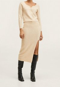 Beżowy sweter dzianinowy z dekoltem w kształcie litery V i trzema guzikami, zestawiony z dopasowaną midi spódnicą z bocznym rozcięciem. Noszony z czarnymi kozaczkami do kolan.