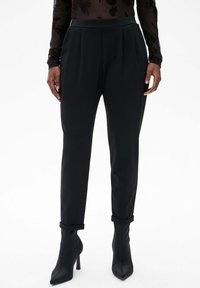 Zwarte op maat gemaakte broeken met plooien, een gladde textuur en taps toelopende pijpen, gecombineerd met zwarte high-heeled enkellaarzen.