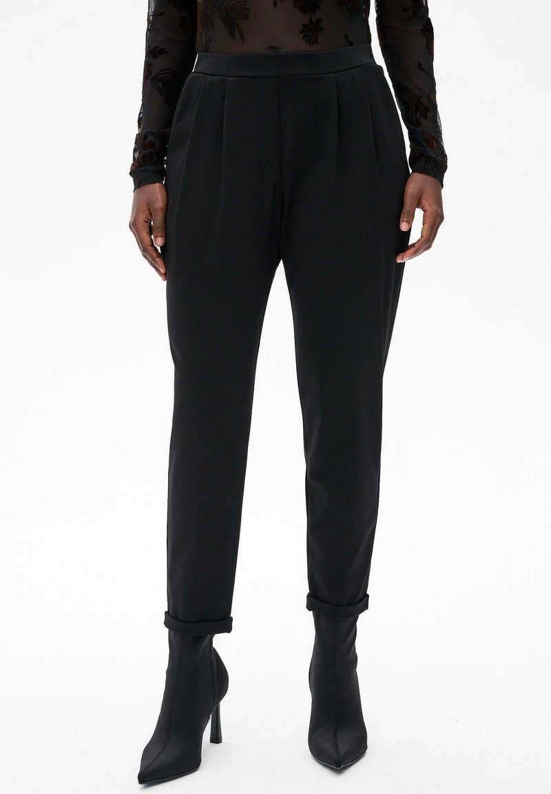 Zwarte op maat gemaakte broeken met plooien, een gladde textuur en taps toelopende pijpen, gecombineerd met zwarte high-heeled enkellaarzen.