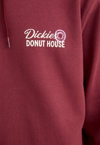 Hanorac maroon cu textul "Dickies Donut House" în alb, însoțit de o grafică cu o gogoașă. Fabricat dintr-un material moale, texturat. Glugă ajustabilă.