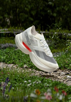 White and gray athletic shoe with red accent, floating above a gravel path surrounded by green plants and small flowers.