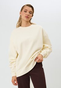 Felpa color crema con scollo rotondo, vestibilità rilassata e polsini a coste. Abbinata a pantaloni a coste di colore marrone scuro.