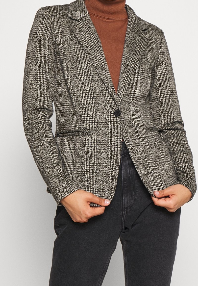 Femme portant un blazer à carreaux noir et blanc à un seul bouton, par-dessus un pull à col roulé couleur rouille et un pantalon noir.