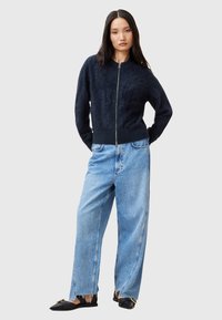 Navy gestrickter Zip-Pullover mit strukturierter Oberfläche, kombiniert mit hellblauen weiten Jeans und flachen schwarzen Schuhen mit goldenen Akzenten.