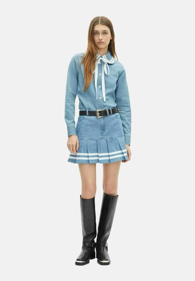 Jonge vrouw in een spijkerblouse met een grote kraagstrik, geplooide spijker mini-rok met witte strepen, een zwarte riem en kniehoge zwarte laarzen.