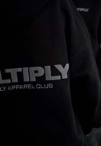 Sort hættegammert med stor grå tekst "LTIPLY" og mindre hvid broderet tekst "LY APPAREL CLUB" på ryggen, tæt på udsigt over stofstrukturen.
