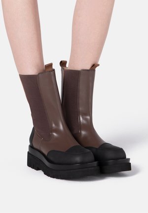 Bottes en cuir marron mi-mollet avec embouts noirs et semelles épaisses noires, portées sur jambes nues sur un fond uni.