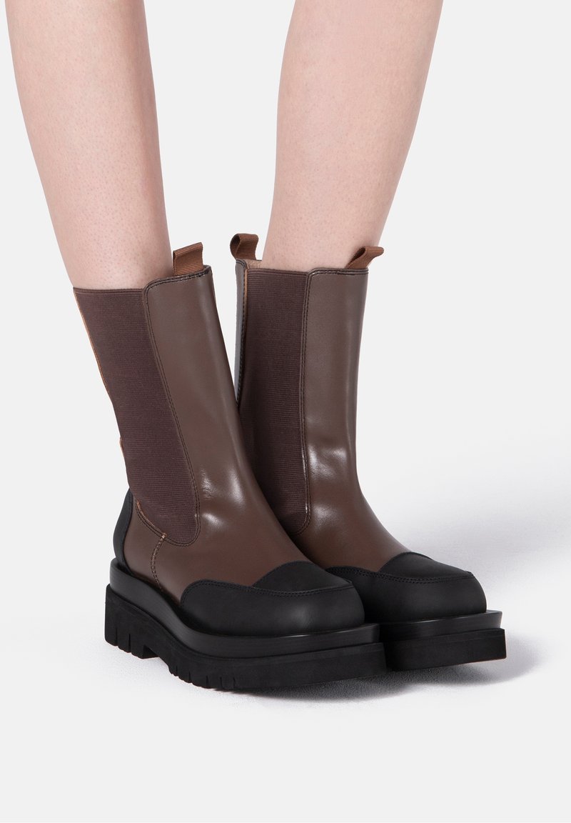 Bottes en cuir marron mi-mollet avec embouts noirs et semelles épaisses noires, portées sur jambes nues sur un fond uni.