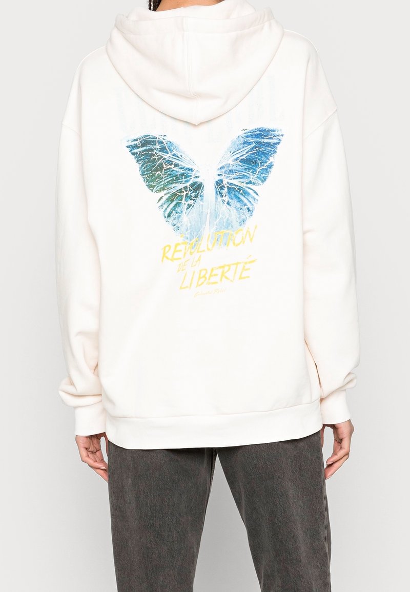 Sweat à capuche blanc avec un grand motif de papillon bleu au dos, orné de textures bleues et vertes, et du texte "RÉVOLUTION DE LA LIBERTÉ."
