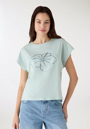 Top a maniche corte verde menta chiaro con una vestibilità ampia, caratterizzato da un disegno floreale con paillettes al centro e scollo rotondo.