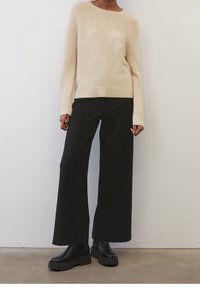 Pull en beige duveteux avec des manches longues et un ourlet côtelé, associé à un pantalon large noir et des bottes noires imposantes. Texture lisse sur l'ensemble.
