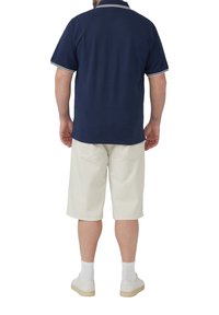 Marineblaues Polo mit weißem Kragen und Ärmelborten, kombiniert mit beigen knielangen Shorts und weißen Sneakern, von hinten betrachtet.