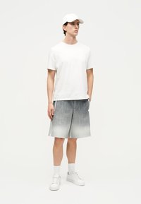 Mladý muž v bielej čiapke, bielom tričku, sivých ombré šortkách, bielych ponožkách a bielych teniskách stojaci pred čistým bielym pozadím.