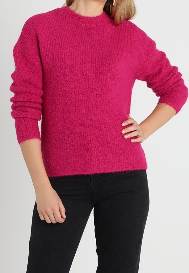 Donna che indossa un maglione peloso di un vivace colore rosa con maniche lunghe e pantaloni neri, in piedi con una mano sulla vita e l'altra rilassata.