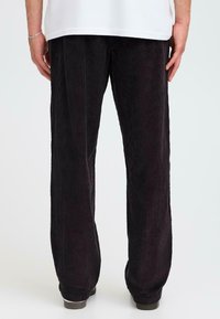 Schwarze Cordhose mit vertikal gerippter Textur, im relaxte Schnitt und leicht ausgestellten Beinen. Der Bund ist mit einem dezenten Detail versehen.