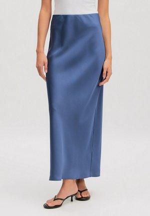 Woman wearing a blue satin midi skirt paired with black heeled thong sandals and a white sleeveless top, standing against a plain background.