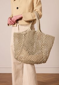 Sac fourre-tout en filet beige clair, fabriqué en matériau tissé, avec deux poignées, un design ouvert et une finition texturée.