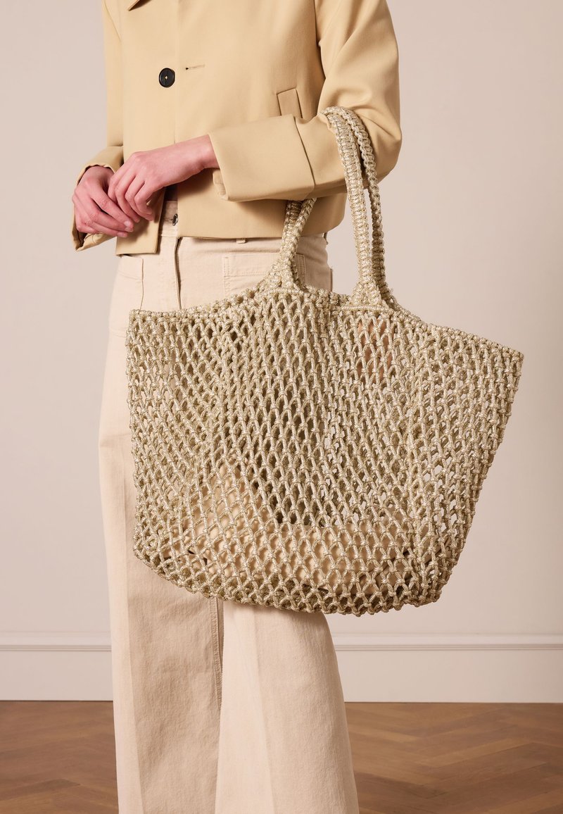 Sac fourre-tout en filet beige clair, fabriqué en matériau tissé, avec deux poignées, un design ouvert et une finition texturée.