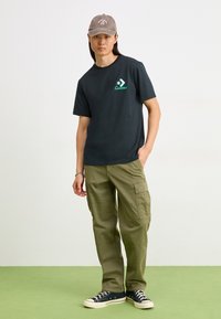 Camiseta de algodón negra con logo turquesa, combinada con pantalones cargo verde oliva y zapatillas de lona azul marino sobre un suelo verde. Sombrero con logo.