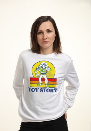 Frau mit braunen Haaren, die ein weißes Langarmshirt mit Buzz Lightyear und Toy Story Schriftzug trägt, vor einem schlichten Hintergrund stehend.