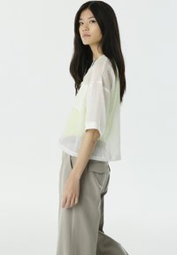 Weißes und hellgrünes transparents Kurzarmpoloshirt, lockere Passform mit verkürzter Silhouette; kombiniert mit hellbeigen Hosen.