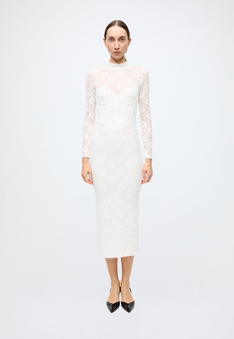 White lace long-sleeve dress with sweetheart neckline, fitted silhouette, and mid-calf length. Black pointed-toe heels complete the look.