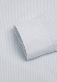Manche de chemise bleu clair avec de fines rayures blanches verticales et coutures visibles sur le poignet, sur un fond de tissu rayé assorti.