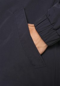 Granatowa kurtka z gładkiej, lekkiej tkaniny; posiada marszczone rękawy oraz boczną kieszeń z czystym wykończeniem krawędzi.